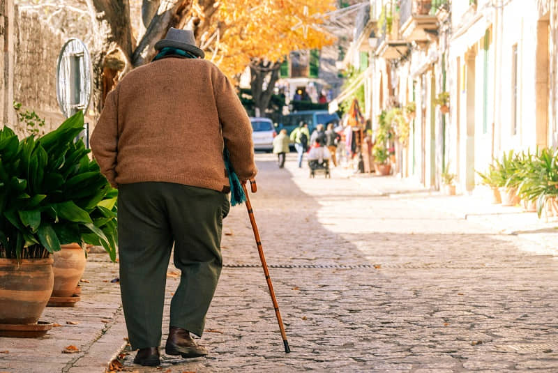 Senior person with walking cane strolls down sunny cobblestone street lined with potted plants and shops