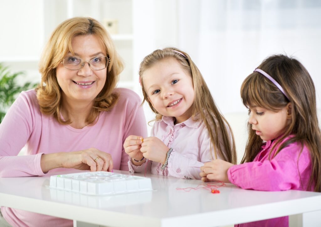 Grandmother and two young girls smiling and making beaded jewelry together at a table, enjoying a fun and creative bonding activity.