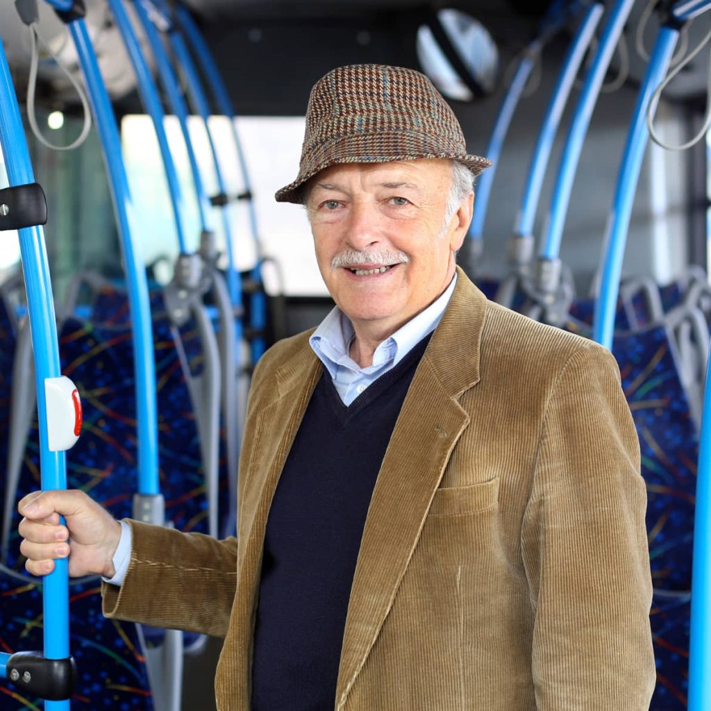 Smiling senior man in tweed jacket and cap riding public bus, holding handrail while standing in the aisle